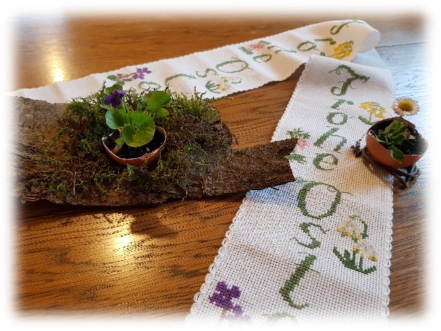 Längliche Tischdecke mit gesticktem Schriftzug 'Frohe Ostern' und Blumenmotiven, daneben ein Stück Baumrinde mit Moos und kleinen Blumentöpfen mit violetten und gelben Blüten.