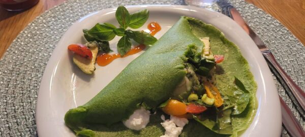 Auf einem weißen Teller liegt ein grüner, gefalteter Pfannkuchen mit Füllung aus Lachs, Salat und kleinen Tomaten, daneben ein Klecks Sauce und einige Basilikumblätter.