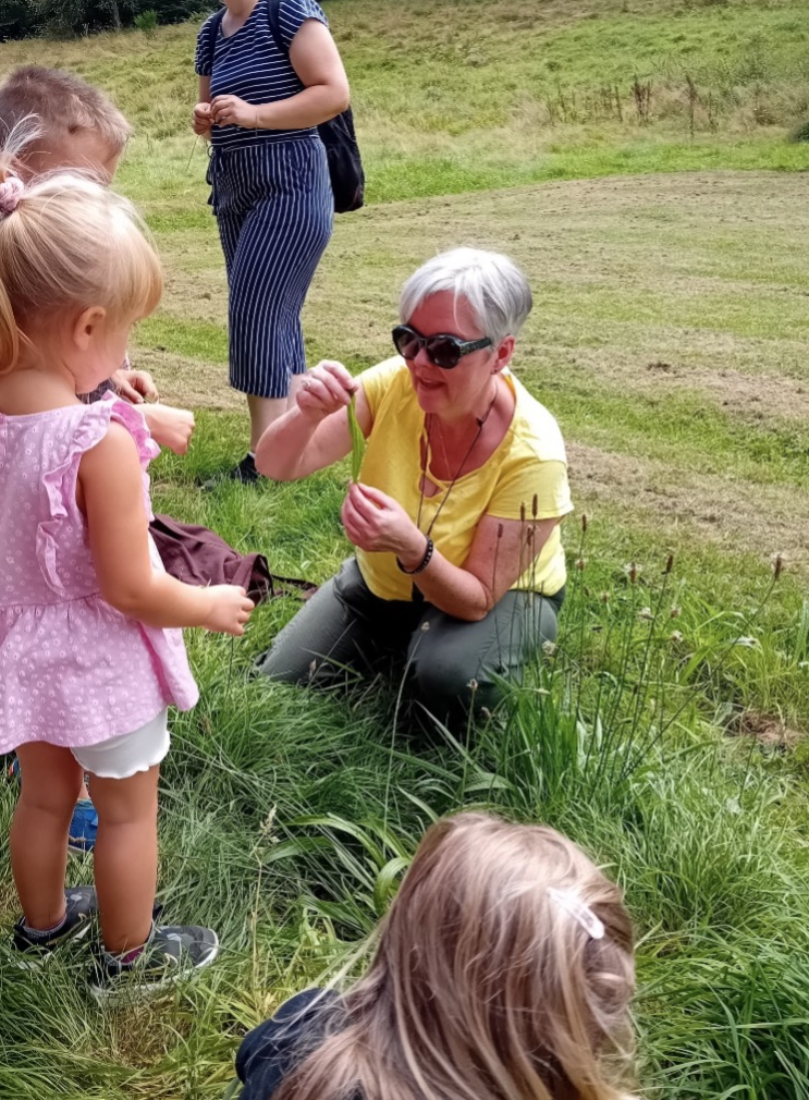 Kräuterfrau Eva kniet im Gras und zeigt Kindern bei einem Kräuterspaziergang der KiTa eine Pflanze