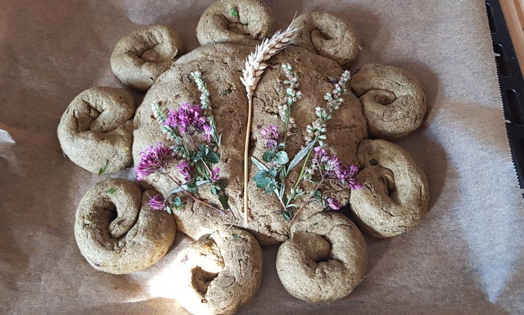 Ein Brotteig zu einer Sonne geformt. Darauf liegen verschiedene bunte Kräuter und eine Getreideähre