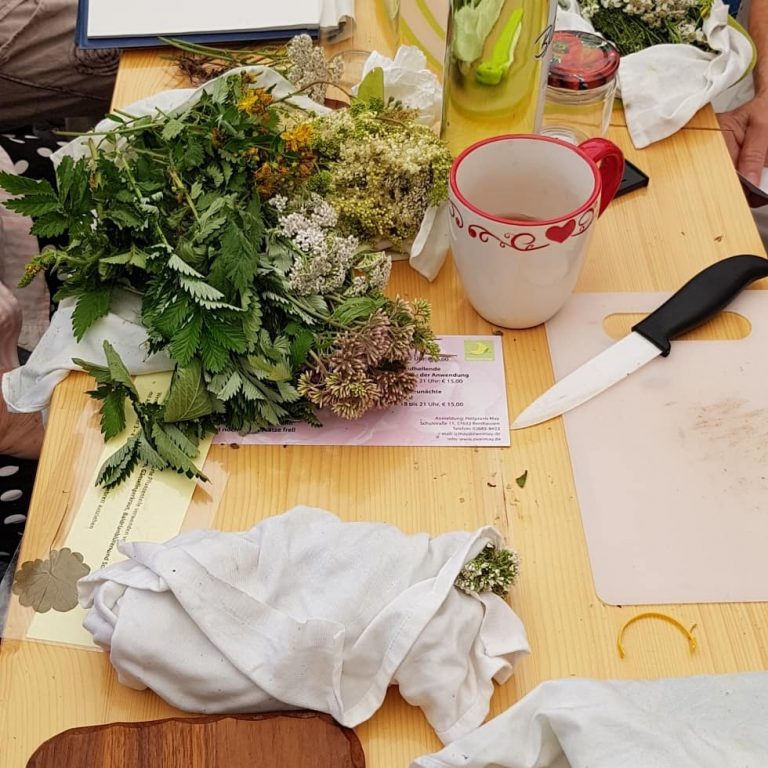 Tisch mit verschiedenen frischen Kräutern, einem Messer, einer Tasse mit roten Herzen und einem Zettel zum Workshop Kräuterwanderung